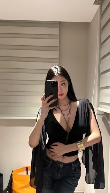 A woman taking a mirror selfie with her phone, wearing a black top and sheer overlay, a gold watch, and layered necklaces.