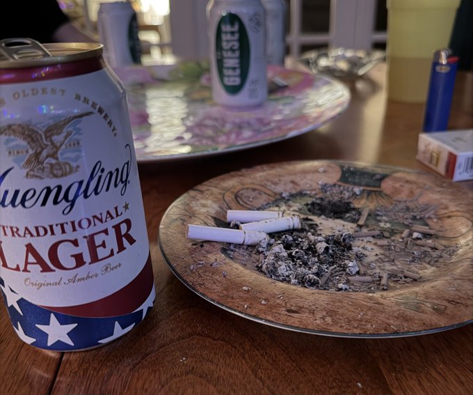 A Yuengling Traditional Lager can sits beside an ashtray overflowing with cigarette butts, with a Genesee can and lighter visible in the background. The scene captures the friend's dad mid-rant about Elon and Optimus, surrounded by the smoking-and-beer-fueled atmosphere the post describes.