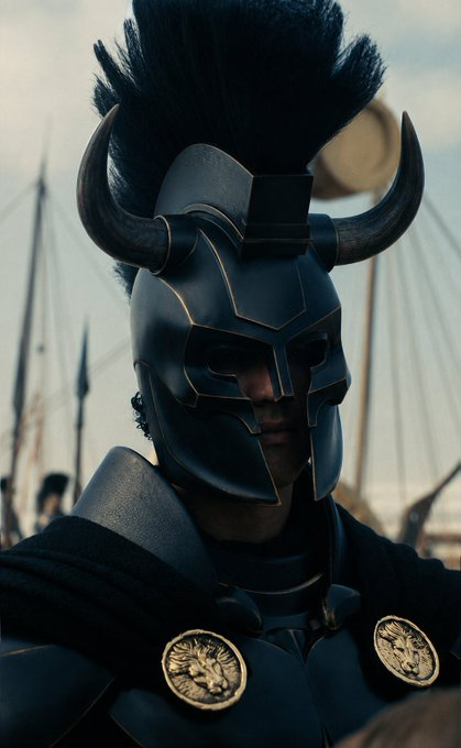 A dramatic portrait of a warrior wearing an ornate black horned helmet with gold accents, standing before spears and a cloudy sky.