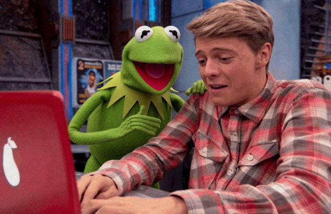 Kermit the Frog and a young man in a plaid shirt look at a red laptop together, from what appears to be the set of a TV show.