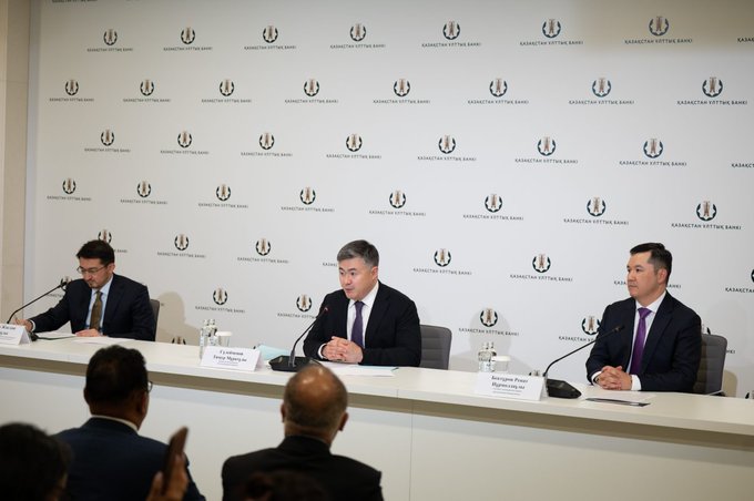 Three men in business suits sit at a panel table against a backdrop displaying "Kazakhstan KTTB Bank" logos, with name placards visible including "Daniyar Yerikovich" and "Daniyarovich Ruslan." The image appears to relate to the accompanying post about Kazakhstan's regulatory developments in cryptocurrency mining, showing officials likely involved in the country's blockchain and digital economy discussions.