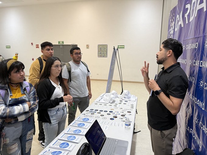 A Cardano representative explains blockchain concepts to students at the FePro 2026 event in Puebla, Mexico, with "CARDANO" branding displayed on a banner beside a table of educational materials.