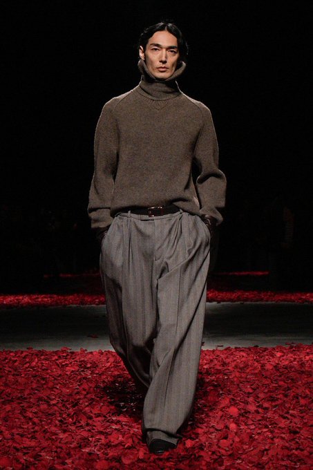 A male model walks a runway covered in red rose petals, wearing a brown turtleneck and loose gray trousers.
