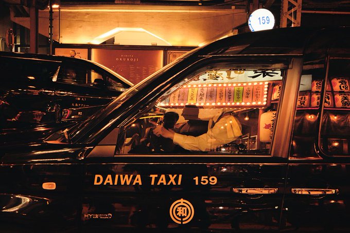 A night scene of a Japanese taxi driver in a black 'DAIWA TAXI 159' cab, wearing a white shirt and cap, with illuminated menus and signs visible through the car windows.