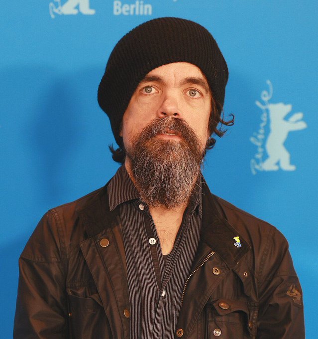 A bearded man wearing a black beanie and dark jacket poses in front of a blue backdrop with "Berlin" and a Berlinale bear logo visible, suggesting a press event at the Berlin International Film Festival.
