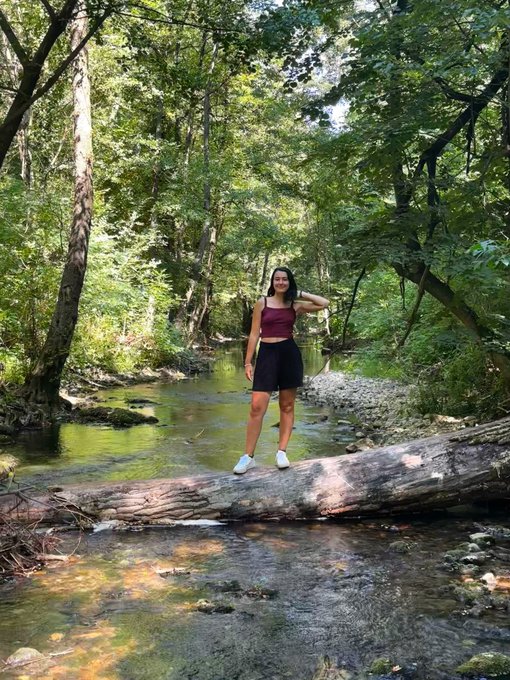 A young woman stands on a fallen log spanning a tranquil forest stream, posing for a photo amid lush greenery. The image complements the caption's mention of her 20-minute nature conservation walks, capturing a peaceful moment that reflects her commitment to environmental activities alongside her crypto achievements.