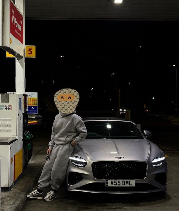A person wearing a gray tracksuit leans against a silver Bentley with the license plate 'V55 BML' at a V-Power gas station at night, their face obscured by a heart-shaped emoji.