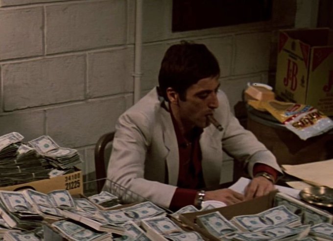 A scene from a film showing a man with a cigar in his mouth sitting at a desk counting stacks of cash, surrounded by bundles of money in boxes and J&B whisky boxes in the background.