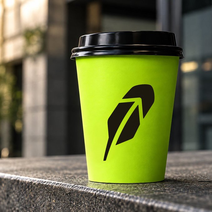 A bright green paper coffee cup with a black lid and a black feather/quill logo, sitting on a stone ledge against a blurred urban background.
