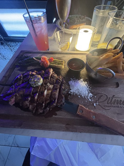 A meal at Pitmaster Steakhouse & Lounge featuring a large steak with butter on a wooden board, served with vegetables, a dipping sauce, fries in a metal basket, salt, and drinks.