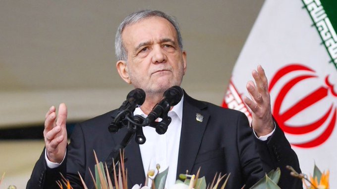 Iranian President Masoud Pezeshkian speaks at a podium with multiple microphones, wearing a dark suit, with the Iranian flag visible behind him.