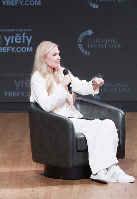 A blonde woman in a white pantsuit sits in a black leather chair on a stage, holding a microphone in front of a backdrop featuring 'YREFY.COM' and 'TURNING POINT USA' logos.