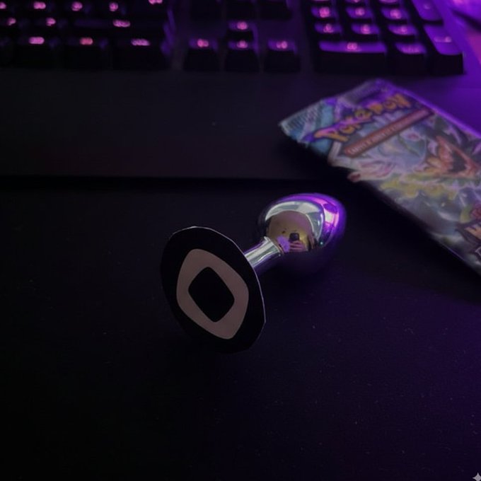A silver butt plug lies on a dark desk mat in front of a backlit gaming keyboard with purple lighting, with a Pokémon trading card pack visible to the right.