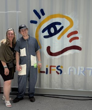 In the image, there are two people standing next to each other against a white wall with colorful graffiti art on it. The person on the left is wearing blue overalls and has their arm around the person on the right who is dressed in black. They both have smiles on their faces as they pose for the photo. 

The post features an image of a face, likely a painting or drawing, with the words "Lifes" written above it. The graffiti art on the wall adds a vibrant and artistic touch to the scene.