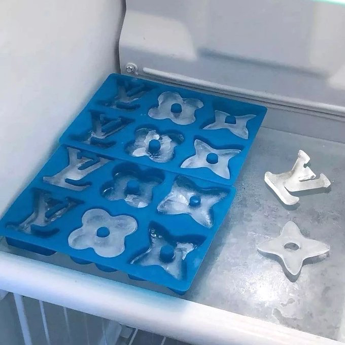 A blue silicone mold sits in what appears to be a freezer, containing partially frozen shapes including stars, flowers, and the letters 'Y' and 'V', with two completed silver-colored frozen shapes resting on the freezer surface beside it.