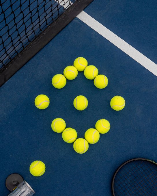 Yellow tennis balls arranged in a circular pattern on a blue tennis court, with a tennis net visible in the corner.