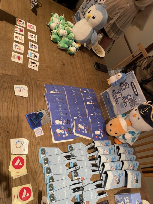 A wooden table displaying an assortment of merchandise including Sanrio character plushies (Piplup, Cinnamoroll, Pochacco), pins, stickers, hair clips, and what appear to be MNet/Avex promotional items for a K-pop idol group.