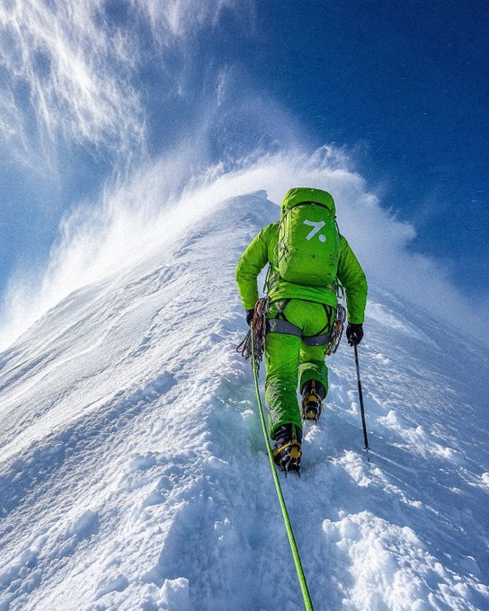 A person in a bright green climbing suit and backpack ascends a steep, snowy mountain ridge under a blue sky, attached to a green rope.