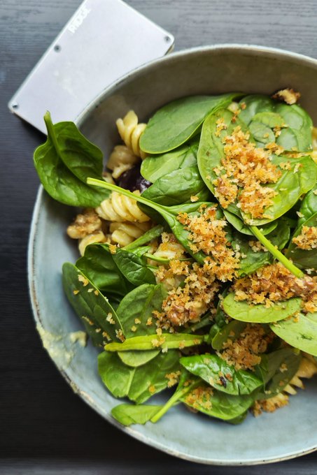 A bowl of spinach salad topped with toasted breadcrumbs and pasta sits next to a metal bench scraper.