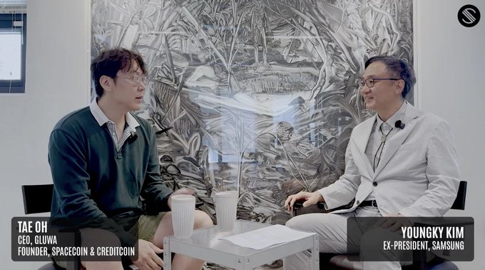 The image features two men sitting at a table with cups of coffee placed between them, engaged in conversation. They are positioned against a wall adorned with an artwork depicting trees and bushes. The background includes text that reads "@Spacecoin", "@Spacecoin", and "#spacecoins". This suggests the image is related to the space industry or cryptocurrency, possibly featuring a discussion about the recent SpaceX IPO.