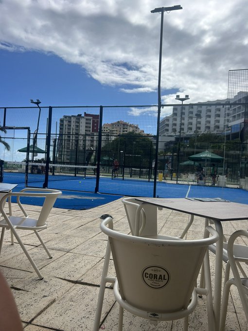 A patio with white chairs and a table bearing a "CORAL" logo overlooks a padel court where players are active, framed by city buildings and a partly cloudy sky — a relaxed setting suggesting a leisure break where one might be asked to share their Solana (SOL) wallet address.