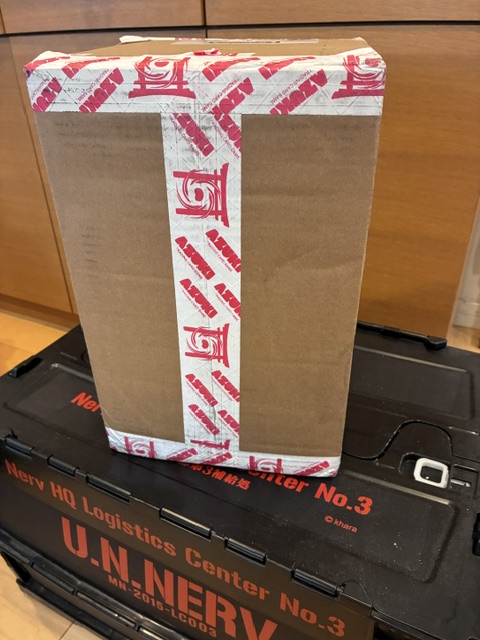 A cardboard box sealed with branded tape sits atop a black storage case marked "Nerv HQ Logistics Center No.3" and "U.N.NERV" with "Trailer No.3" and the code "MR-2016-LCD02," representing a newly arrived shipment from Azuki TCG to a collector in Japan. The image captures the excitement of the unboxing moment, tying into the post's celebration of the growing global community of collectors connected through the brand.