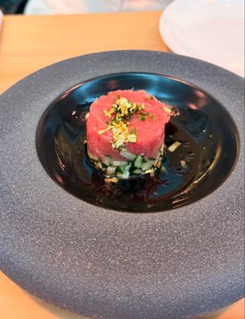 A plated appetizer featuring a cylindrical pink/red tartare (likely tuna or watermelon) garnished with chopped pistachios, served on a dark plate with a base layer of diced vegetables.