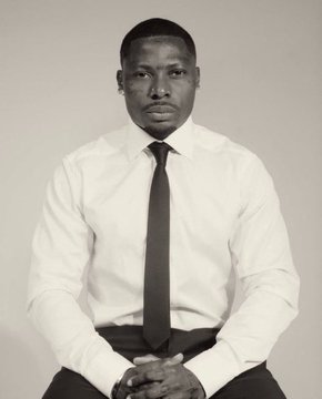 The image features a man sitting down, wearing a white shirt and black tie. He has his hands clasped together in front of him, giving off an air of confidence or contemplation. The photo appears to be in black and white, which adds a timeless quality to the scene. There is no text present in the image, but it seems that the man's attire might have been inspired by the post "solana last year solana this year".