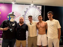 Four men pose for a photo at what appears to be a gaming event, with three wearing casual t-shirts and one wearing a black shirt and a dark mask.