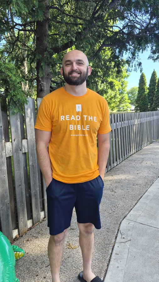 A smiling bald man with a beard stands outdoors wearing an orange t-shirt that reads 'READ THE BIBLE orange.bible.ch' along with navy shorts.