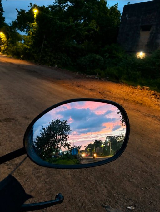 A photograph taken at dusk showing a motorcycle's side mirror reflecting a colorful pink and blue sunset sky with silhouetted trees and a road with headlights, while the actual road and surrounding dark vegetation are visible in the background.