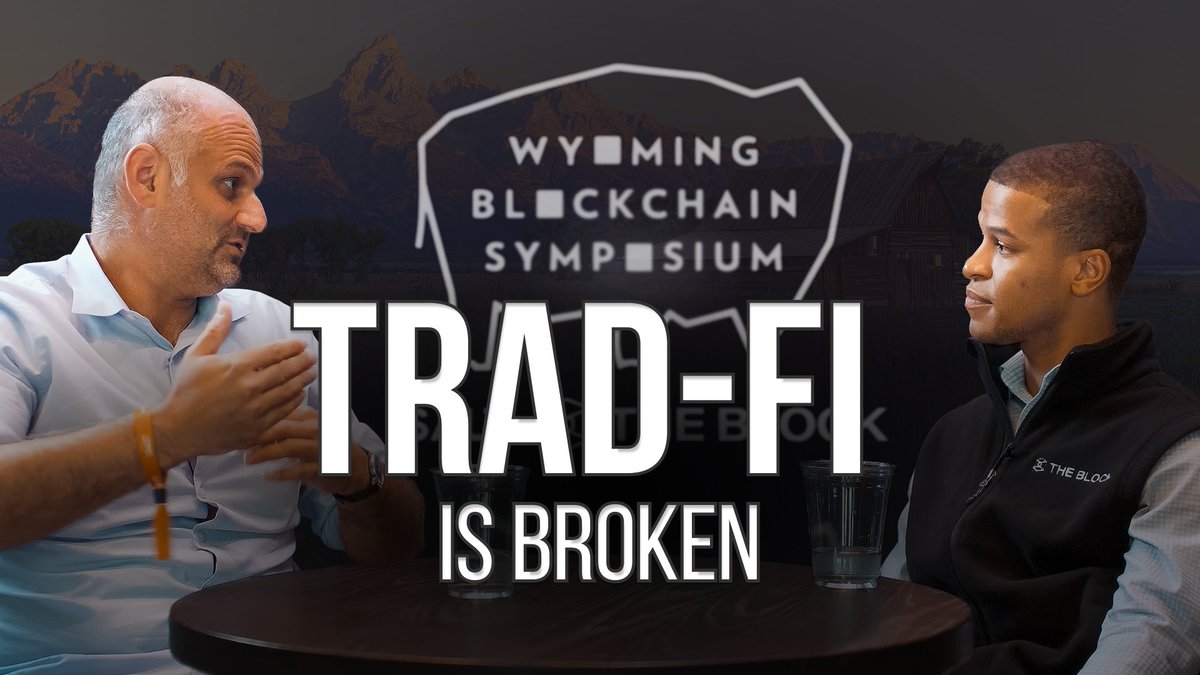 Two men sit at a table in conversation with the text 'TRAD-FI IS BROKEN' overlaid, set against a backdrop reading 'WYOMING BLOCKCHAIN SYMPOSIUM' over a mountain scene.