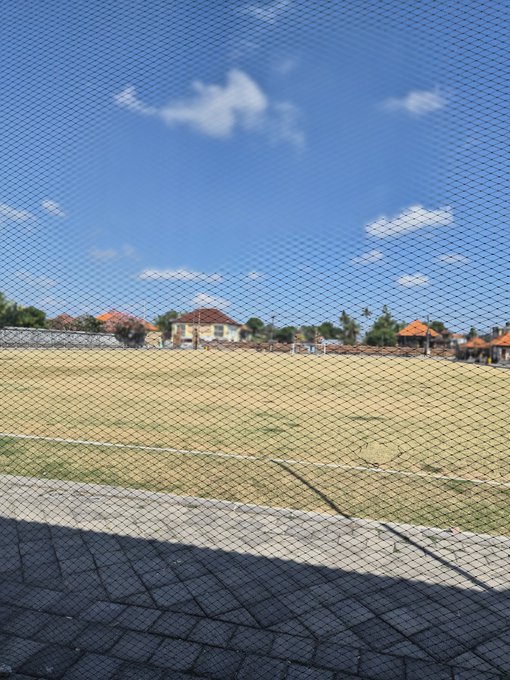 A view through a chain-link fence of a grassy sports field under a blue sky with clouds, with houses visible in the background.