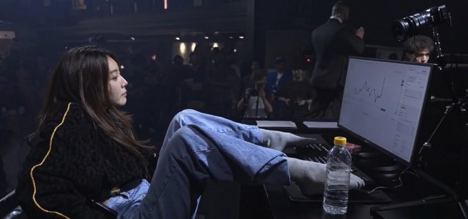 A young woman with long dark hair sits with her feet up in front of a computer monitor displaying a chart, holding a water bottle, in what appears to be an esports or gaming event setting.