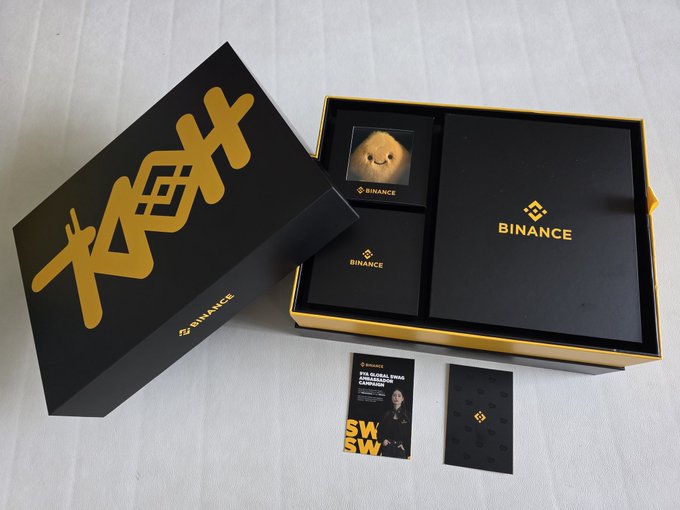 The image shows an open black and gold Binance 9th anniversary gift box containing a plush toy, branded cards, a "SWAG SUCCESS" voucher featuring what appears to be CZ (Changpeng Zhao), and a Binance-branded item, all displayed on a white surface—celebrating Binance's 9th birthday as mentioned in the post.