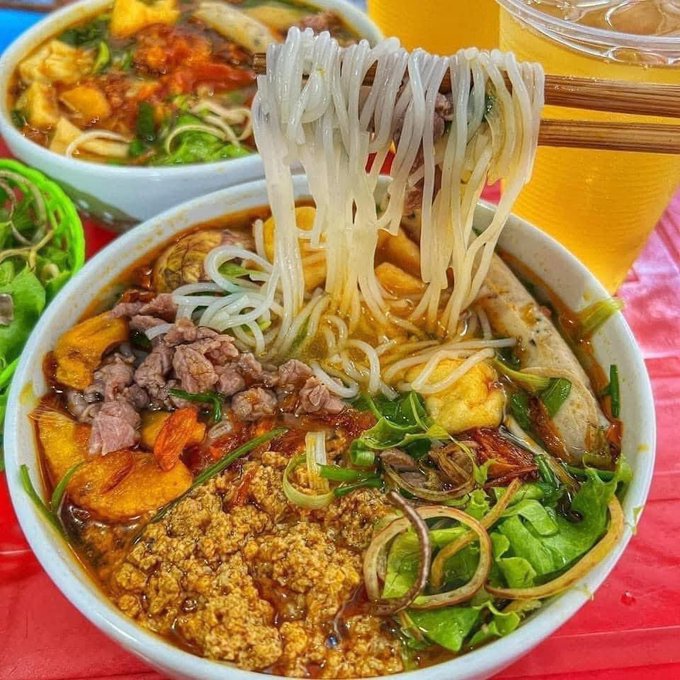 A close-up of a bowl of Vietnamese-style noodle soup with rice noodles being lifted by chopsticks, accompanied by a glass of iced tea.
