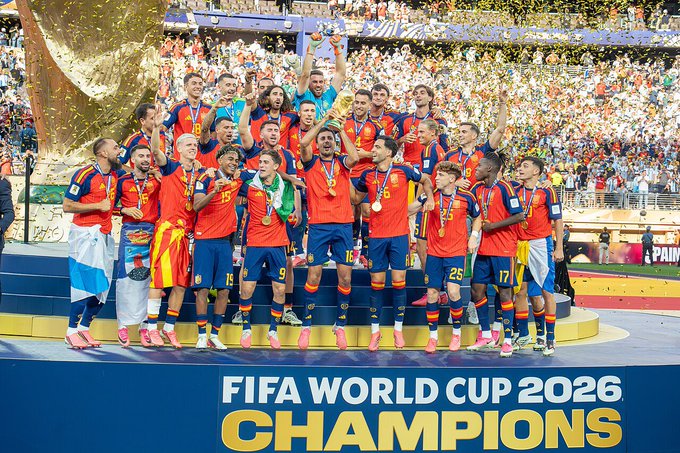 A soccer team in red and blue uniforms celebrates on a podium with a trophy, surrounded by confetti, with text reading 'FIFA WORLD CUP 2026 CHAMPIONS'.