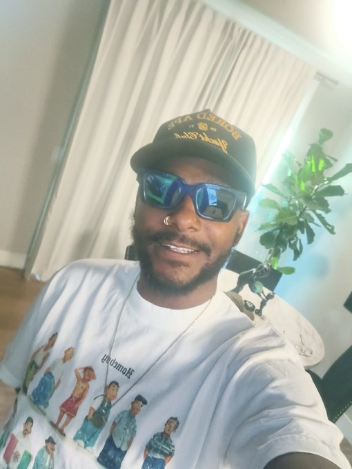 A man wearing a "For the Kids" snapback cap, blue reflective sunglasses, and a white Stüssy graphic t-shirt with character prints smiles for a selfie indoors, with a plant and curtains visible behind him. The image serves as his Day 560 selfie in a series supporting ApeCoin ($APE) until it hits an all-time high, with the "Blessings ApE" caption reflecting his hopeful crypto community spirit.