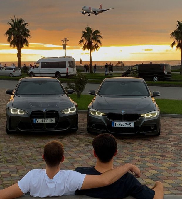 Two BMW sedans are parked on a brick driveway as two people watch an airplane fly overhead during a sunset, with palm trees lining the background.