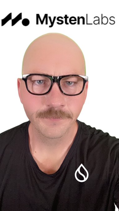 A bald man with glasses and a mustache wearing a black shirt with a Sui logo, with 'MystenLabs' branding visible at the top.