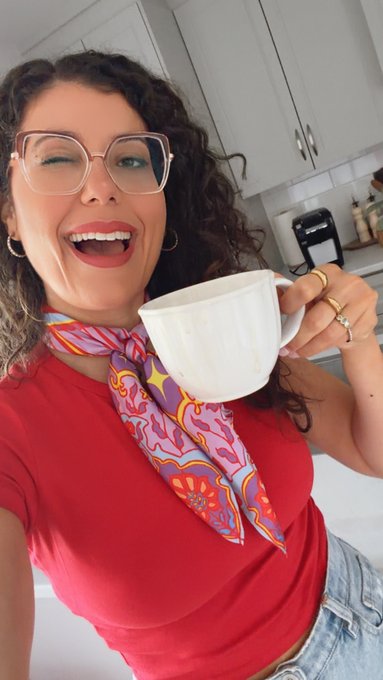 A cheerful woman with curly hair and glasses winking while holding a large white teacup, wearing a red top and a colorful patterned scarf in a kitchen setting.