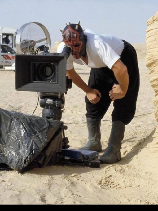A person wearing a Darth Maul mask and boots operates a professional film camera in a desert setting, with a vehicle visible in the background.