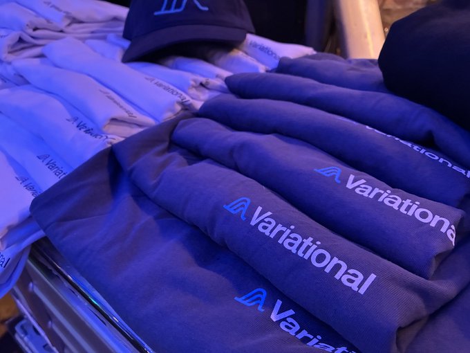 A stack of folded t-shirts and a cap branded with the 'Variational' logo sit on a table, illuminated by blue event lighting.