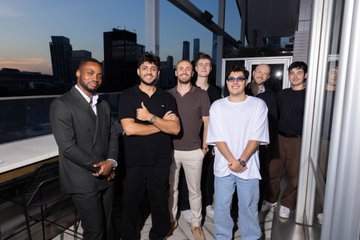 The image features a group of six men posing for a photo on an outdoor balcony or terrace. They are all dressed up, wearing suits and ties, which suggests that they might be attending a formal event or gathering. The backdrop includes buildings, indicating that this location is in New York City. The men appear to be enjoying themselves as they pose together, creating a sense of camaraderie among the group.