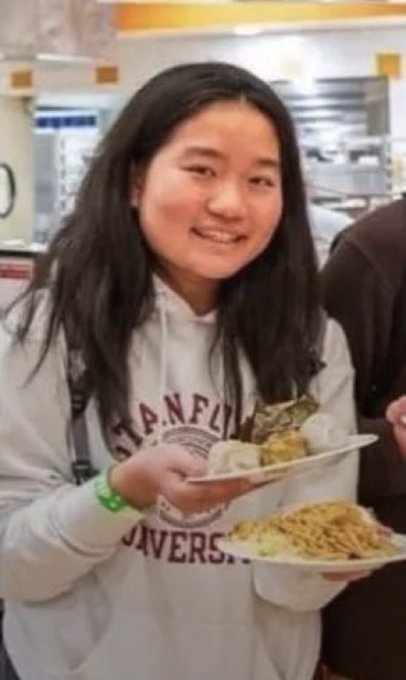 The image shows a smiling young woman with long dark hair wearing a white "Stanford University" hoodie, holding two plates of food (one appears to be rice with greens and another with a noodle dish) in what looks like a cafeteria setting. The post's joke about "looksmaxxed" plays on the humorous contrast between her casual appearance while carrying food and the exaggerated implication of aesthetic enhancement.