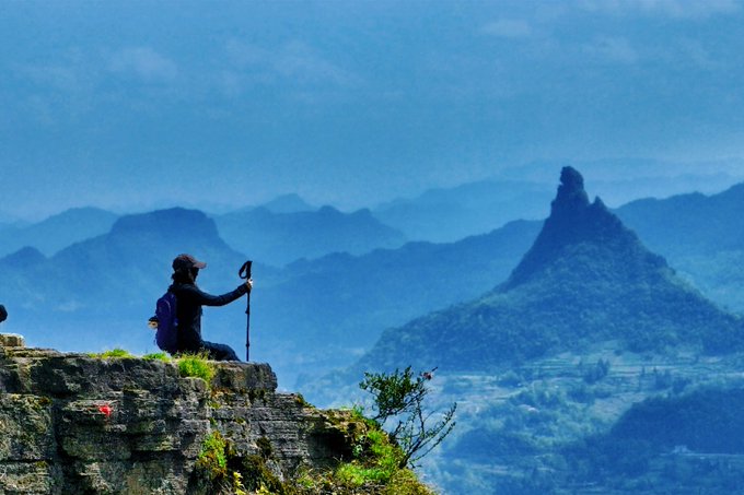 A lone hiker wearing a cap and backpack sits on a rocky cliff edge holding a trekking pole, gazing out over misty blue mountain ranges with a prominent jagged peak in the distance.