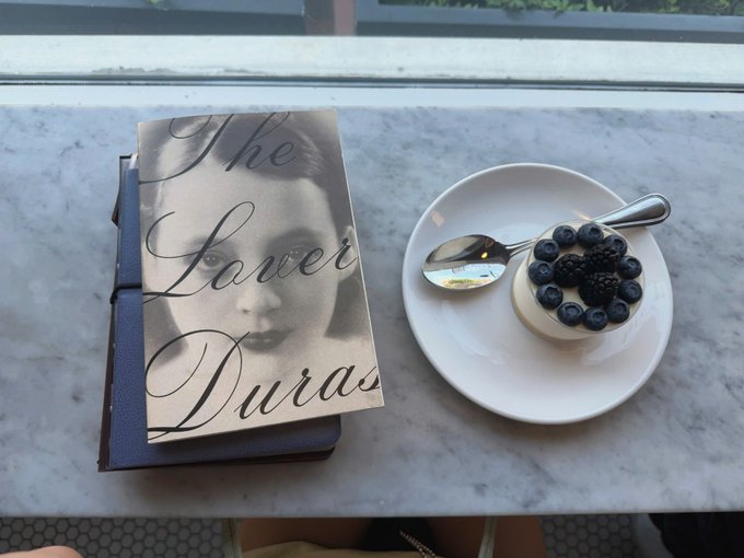 A book titled 'The Lover' by Duras rests on a marble table beside a small dessert topped with blueberries and blackberries on a white plate with two spoons.