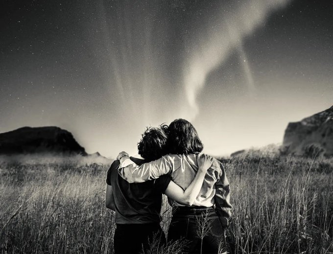 A black-and-white photo of two people standing with their arms around each other in a grassy field, looking up at a starry night sky with wispy cloud formations.
