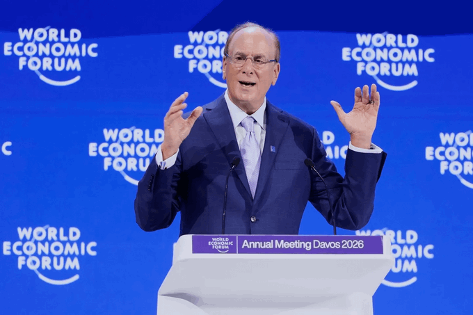 A photograph of a man in a navy suit and light purple tie speaking at a podium during the World Economic Forum Annual Meeting Davos 2026, gesturing with both hands raised.