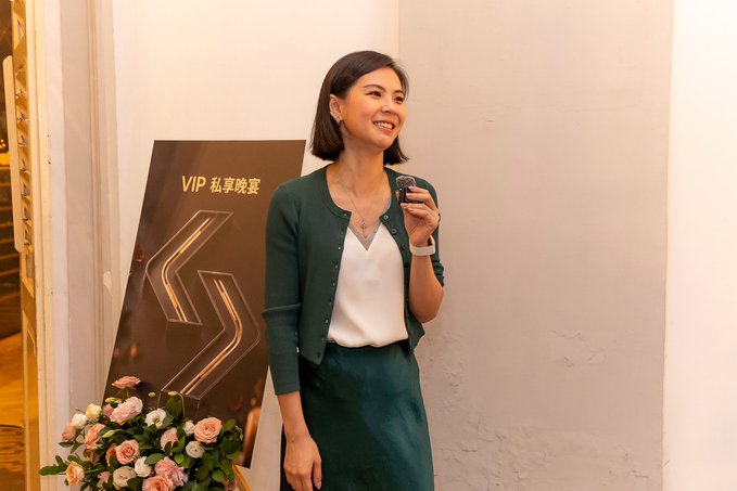 A smiling woman in a green cardigan and white top stands beside a sign reading 'VIP 私享晚宴' (VIP Private Dinner), holding a small recording device.